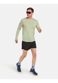 Hellgrünes Langarmhemd und schwarze Shorts, kombiniert mit blauen Knöchelsocken und dunklen Sportschuhen. Die Person trägt dunkle Sonnenbrillen.