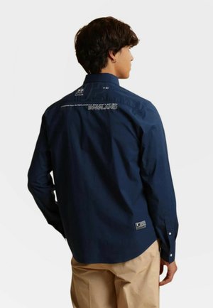 Man met rug naar de camera, draagt een navyblauw overhemd met knopen met de tekst "England" op de bovenrug en beige broek tegen een witte achtergrond.