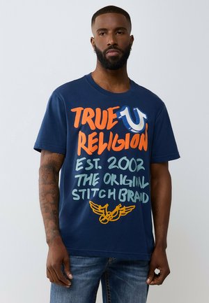 Mann mit Bart und Tattoos trägt ein dunkelblaues True Religion T-Shirt mit großem orangefarbenem, hellblauem und gelbem Text und Symbolen, kombiniert mit blauen Jeans.
