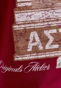 Bordo audums ar nolietu tekstūru, kurā ir balti un brūni drukāti teksti, tostarp grieķu burti un "Originals Atelier."