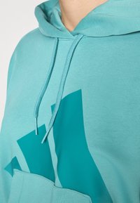 Tirkizna kapuljača s kengur džepom, kapuljačom s vezicama i velikim teal logotipom. Mekani pamučni materijal s glatkom teksturom.