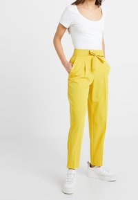 Gele high-waisted broek met een strikceintuur, geplooide voorkant en zijzakken, gecombineerd met een aansluitend wit shirt met korte mouwen en witte sneakers.