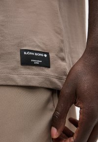 Κοντινό πλάνο του χεριού ενός ατόμου κοντά σε μπεζ ρούχο με μια μαύρη ετικέτα που γράφει «BJÖRN BORG STOCKHOLM 1956» ραμμένη στο τελείωμα του πουκάμισου.