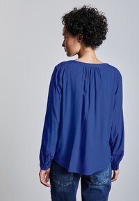 Blusa azul de manga larga con escote redondeado y detalle fruncido en la parte trasera. Tejido suave con una textura lisa, combinada con unos jeans de mezclilla.