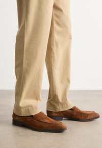 Loafers din suede maro cu un finisaj texturat și o bandă distinctă pe partea superioară, asortați cu pantaloni bej deschis.