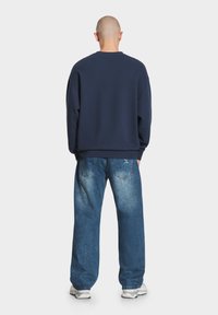 Granatowy sweatshirt z okrągłym dekoltem i obniżonymi ramionami, w połączeniu z luźnymi niebieskimi dżinsami o wyblakłym wykończeniu i dwoma tylnymi kieszeniami.