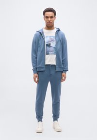 Νέος άνδρας που φοράει μπλε φούτερ με φερμουάρ, αντίστοιχο παντελόνι φόρμας, λευκά αθλητικά παπούτσια και λευκό T-shirt με γραφικό κύμα και το κείμενο «HUGO».