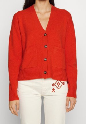 Femme portant un cardigan rouge vif boutonné avec deux poches devant et un pantalon blanc avec une broderie géométrique rouge sur la poche.
