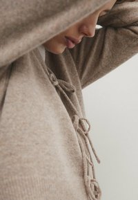 Beige neulepaita, jossa on teksturoitu pinta ja nyöritykset kaula-aukossa. Kangas vaikuttaa pehmeältä ja mukavalta.