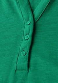 Zelený bavlněný top s výstřihem ve tvaru V, který má pět shodných knoflíků a vertikální žebrovanou texturu. Švy podél okrajů.