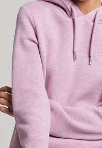 Sudadera de color rosa claro hecha de material suave, con un escote en forma de V, cordones ajustables y un pequeño logo bordado en el pecho.