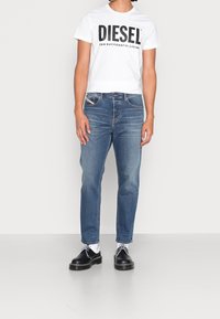 Vit t-shirt med en djärv svart "DIESEL"-logotyp, blå denimjeans och svarta skor med vita detaljer. Avslappnad passform och medelmörka jeans.