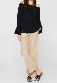 Off-the-shoulder navyblå top med struktureret stof og klokkeærmer, parret med lys beige ternede brede bukser og sorte sandaler.