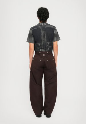 Homme avec des cheveux bouclés portant un T-shirt à motifs noir et gris et un pantalon large marron foncé, se tenant de dos sur un fond blanc.