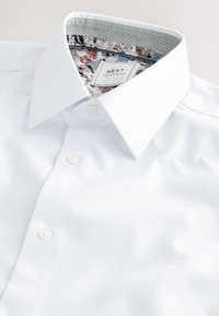 Camicia bianca in tessuto strutturato con colletto a punta, bottoni perlati e dettaglio floreale all'interno del colletto.