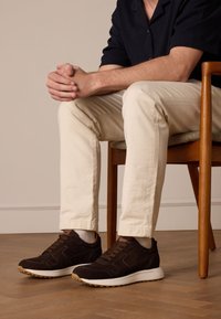 Bruin suede sneakers met witte zolen en bruine veters, gedragen met lichte broek, zittend op een houten stoel.