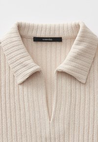 Hellbeige gestrickter Polo-Pullover mit Rippstruktur, einem V-Ausschnitt und einem Etikett mit der Aufschrift "irgendwann".