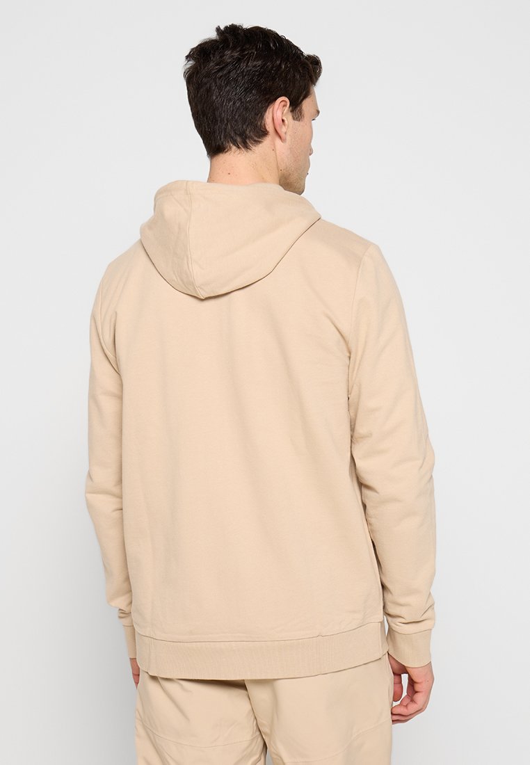 Homme portant un sweatshirt à capuche beige et un pantalon assorti, se tenant de dos contre un fond clair uni.