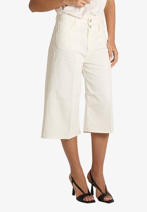 Pantaloni bianchi a vita alta, gamba larga e corta, indossati con sandali neri aperti con tacco alto su una persona in piedi.