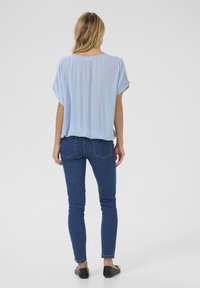 Haut bleu clair, ample, avec des manches courtes retroussées, associé à un jean skinny bleu foncé et des chaussures plates noires. Texture douce, silhouette décontractée.