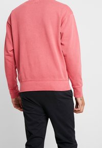 Sweatshirt en coton rose avec manches longues, poignets et ourlet côtelés, présentant une coupe décontractée. La vue arrière montre une texture lisse et sans logos visibles.