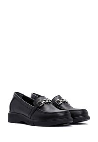 Scarpe slip-on in pelle nera con un design curvo, caratterizzate da un'accento in catena metallica sulla parte superiore e una robusta suola in gomma nera.