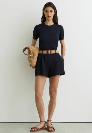 Femeie îmbrăcată cu top cu mâneci scurte bleu închis și pantaloni scurți, cu curea bej, sandale maro, ținând o geantă țesută din paie în fața unui perete alb.