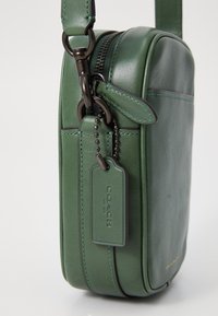 Borsa a tracolla in pelle verde con chiusura a zip, texture liscia e un'etichetta. Presenta una tracolla removibile e dettagli di cucitura discreti.