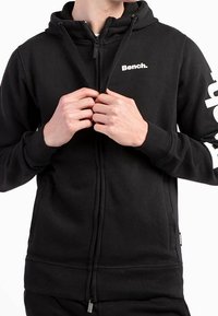 Sudadera con cremallera en negro hecha de un tejido suave. Presenta un logo en el lado izquierdo del pecho y detalles impresos en las mangas. Incluye una capucha con cordón.