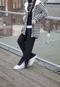 Cappotto a fantasia houndstooth bianco e nero con ampio colletto di pizzo bianco, abbinato a pantaloni neri e sneakers bianche. Sfondo in legno.