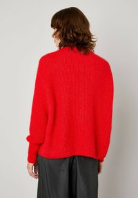 Rdeča pletena jopica z ohlapnim krojem, žametenim videzom in spuštenimi rameni. Ima dolge rokave in okroglo izreza, v kombinaciji s črnimi hlačami.