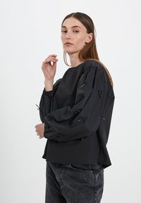 Top nero con maniche a sbuffo e scollatura rotonda, decorato con piccoli fiocchi sulle maniche. Realizzato in un tessuto morbido, abbinato a jeans scuri.
