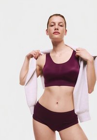 Vinrød sports-bh med rund halsudskæring og sømløs design, kombineret med matchende vinrøde boyshorts. Modellen holder en lysegrå sweatshirt.