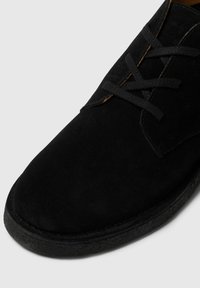 Zwarte suède schoen met een ronde neus, voorzien van zwarte veters en een gestructureerde rubberen zool. Minimalistisch ontwerp zonder extra versieringen.