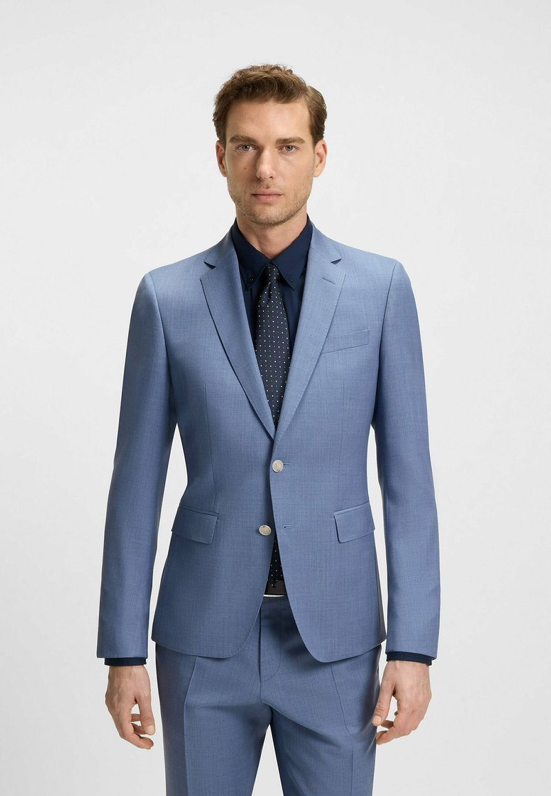 Jasnoniebieska marynarka wykonana z gładkiego materiału, z dwoma przednimi kieszeniami i srebrnymi guzikami, zestawiona z granatową koszulą i krawatem w kropki. Dopasowany krój.