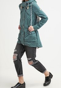 Turkusowy płaszcz przeciwdeszczowy w kropki z kapturem, zapinany na troczki, z dwoma przednimi kieszeniami, zestawiony z postarzanymi czarnymi dżinsami i czarnymi butami.