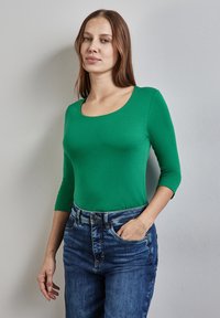 Vihreä, vartalonmyötäinen pitkähihais laukku, jossa on syvä kaula-aukko, valmistettu sileästä kankaasta. Yhdistetty korkeavyötäröisiin, keskimustan sinisiin farkkuihin.