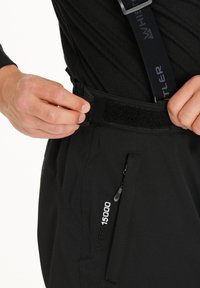 Händer justerar kardborrebandet på midjan på svarta utebyxor med en dragkedjeficka på sidan och märkesförsedda hängslen.