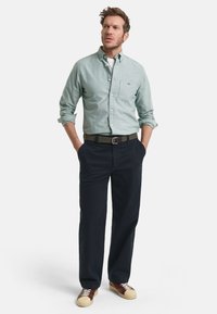Lichtgroene button-up shirt met borstzak, donkerblauwe broek, olijfgroene riem en bruine sneakers met een beige zool, in staande pose.