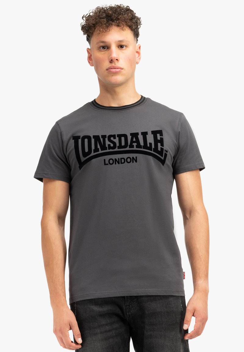 Pelēks kokvilnas T-krekls ar īsām piedurknēm, kas rotāta ar melnu treknu "LONSDALE LONDON" grafiku uz krūts un apaļu kakla izgriezumu.