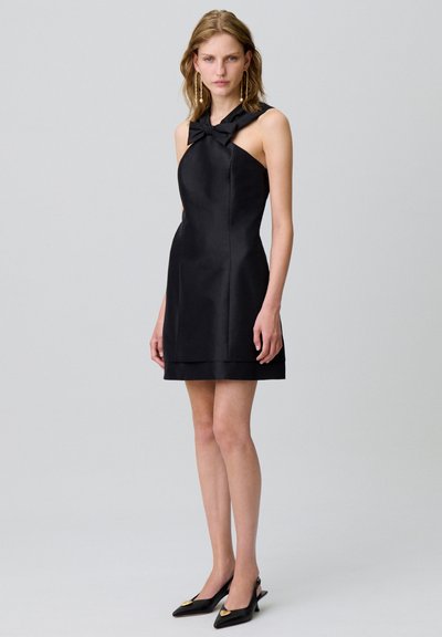 Mini abito in satin nero con scollo a halter e dettaglio a fiocco. Tessuto morbido, silhouette aderente e orlo stratificato. Abbinato a tacchi neri a punta.