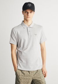 Γκρίζο πόλο μπλουζάκι με γιακά και κοντά μανίκια, που διαθέτει μικρό κεντημένο λογότυπο. Το μοντέλο φοράει ένα μαύρο καπέλο.