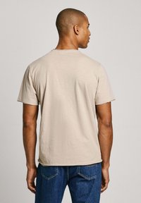 Camiseta de algodón beige con mangas cortas, cuello redondo y corte relajado. Textura suave, diseño liso, combinada con pantalones vaqueros azules.