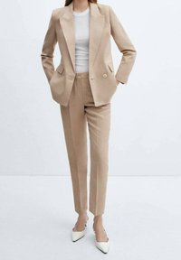 Costume beige sur mesure comprenant un blazer croisé avec de larges revers et des poches à rabat, accompagné d'un pantalon droit et de talons blancs.