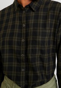 Chemise en flanelle à carreaux verts et noirs avec un col boutonné, une poche avant et des boutons noirs. Texture douce et coupe décontractée.