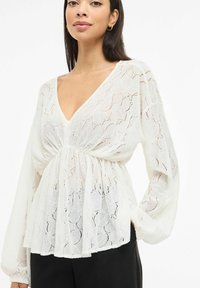Biała, bluzka z dekoltem w kształcie litery V, z fakturowanym wzorem koronkowym, długimi rękawami i marszczonym pasem, zestawiona z czarnymi spodniami.