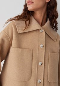Veste en mélange de laine beige avec un col rabattu, col côtelé, fermeture à boutons sur le devant et deux grandes poches plaquées. Texture lisse et design structuré.