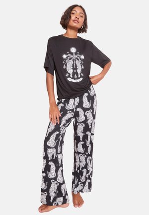 Nainen pukeutunut mustaan löysään t-paitaan, jossa on "Into the Wild" -kuvio, ja siihen sopiviin mustiin housuihin, joissa on valkoisia leopardikuvioita, seisoo paljain jaloin.