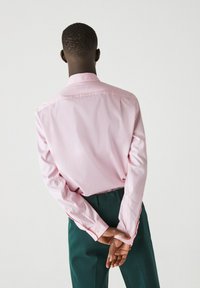 Camicia rosa chiaro a maniche lunghe con colletto button-down e maniche lunghe, abbinata a pantaloni verde scuro. Tessuto morbido, vestibilità casual.
