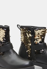 Botas negras hasta el tobillo con parte superior de lentejuelas doradas y negras, suela plana, cremallera lateral y detalle de correa de velcro. Base de cuero suave y acentos texturizados.
